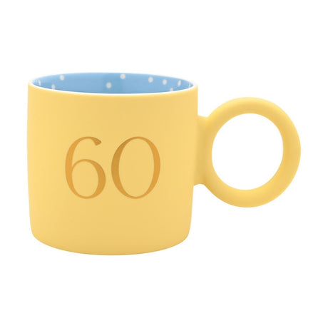 Matt porcelain mug in pastel yellow with a gold "60" print, blue polka-dot interior, and a chunky round ring handle, shown on a plain white background.