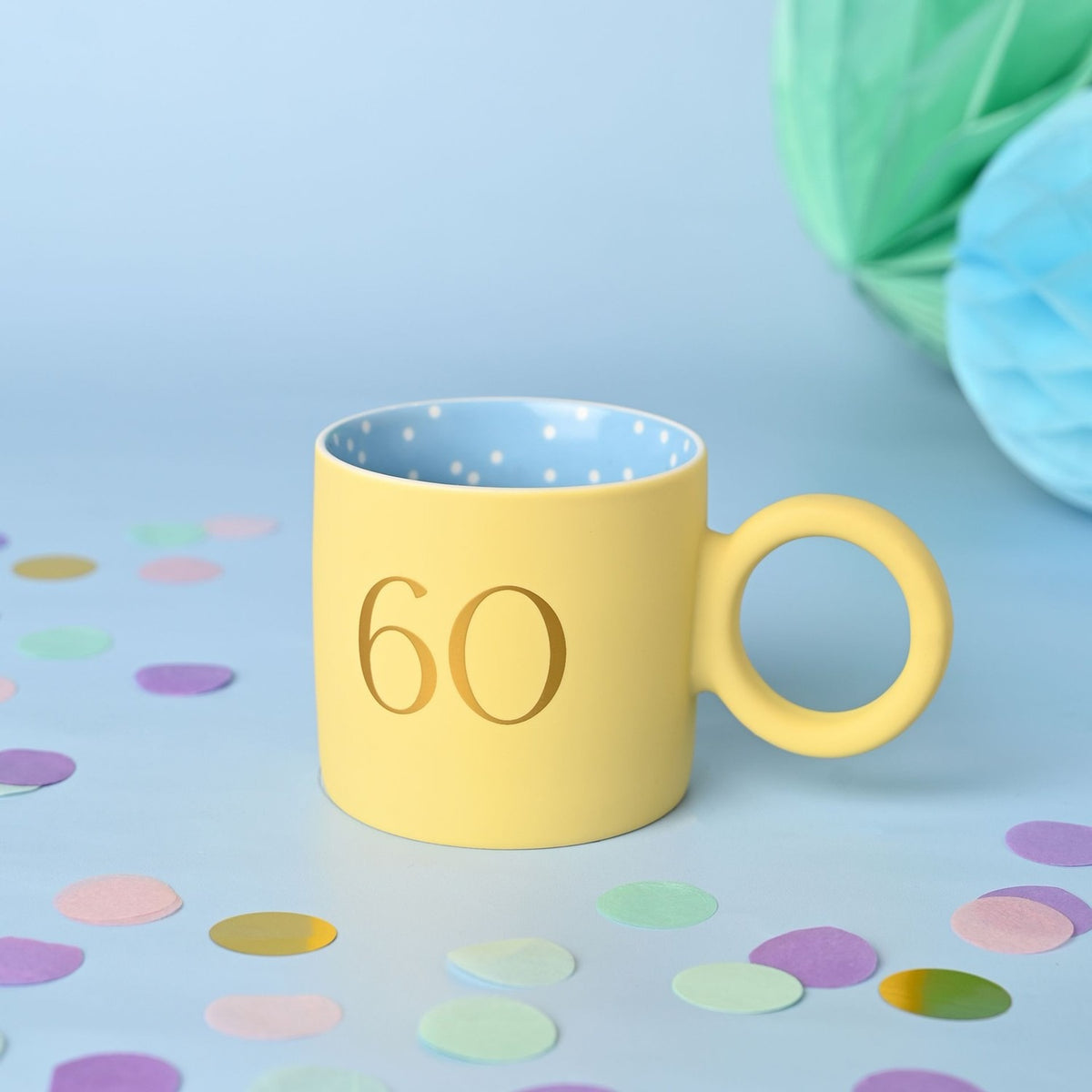 Matt porcelain mug in pastel yellow with a gold "60" print, round ring-shaped handle, and a blue polka-dot interior, set against a light blue background with confetti.