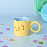Matt porcelain mug in pastel yellow with a gold "60" print, round ring-shaped handle, and a blue polka-dot interior, set against a light blue background with confetti.