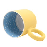 Matt porcelain mug in pastel yellow with a chunky round ring handle, tipped on its side to show the blue, white polka-dot interior, on a plain white background.