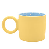 Matt porcelain mug in pastel yellow with a chunky round ring handle, featuring a blue interior scattered with white spots, shown on a plain white background.