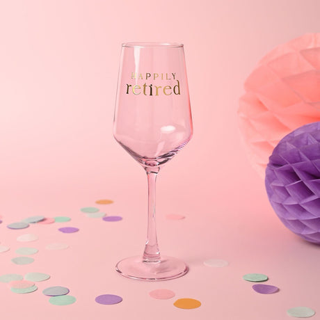 Pale pink stemmed wine glass with gold foil "HAPPILY retired" lettering, shown on a pink background with scattered confetti and honeycomb pom-poms.