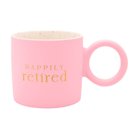 Matt pink porcelain mug with "HAPPILY retired" in gold lettering and a chunky round ring handle, speckled cream interior, shown on a white background.