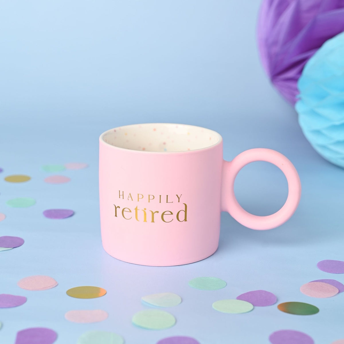 Matt pink porcelain mug with "HAPPILY retired" in gold lettering, a round ring handle, and a speckled cream interior, styled on a blue background with confetti.