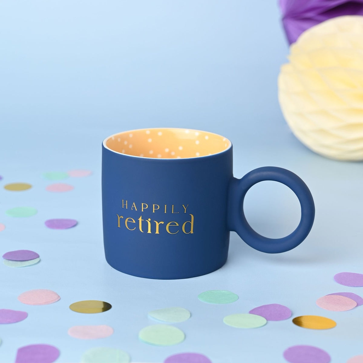Navy matt porcelain mug with "Happily retired" in gold lettering, round navy ring handle, and a yellow interior dotted with small white spots, set against a pale blue background with confetti.