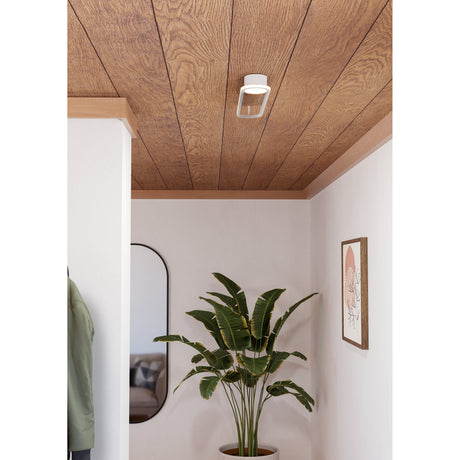Collecorvino LED flush ceiling light in white with a geometric angular shade, shown mounted on a wood-panelled hallway ceiling above a plant and framed wall art.