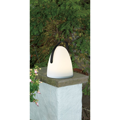 Collirados LED Table Lamp in white plastic with a bell-shaped shade and black arched carry handle, illuminated on a stone pillar against a leafy green backdrop.