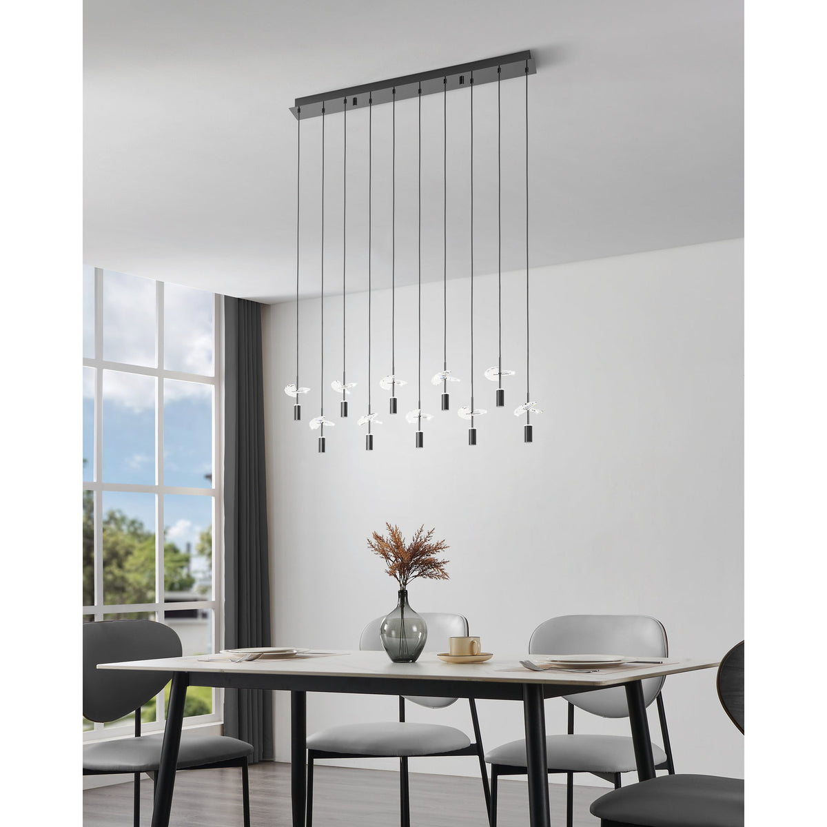 Comareira ten-light linear pendant in black nickel with a long steel canopy and clear faceted glass leaf details, shown hanging above a dining table in a modern living space.