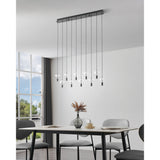 Comareira ten-light linear pendant in black nickel with a long steel canopy and clear faceted glass leaf details, shown hanging above a dining table in a modern living space.