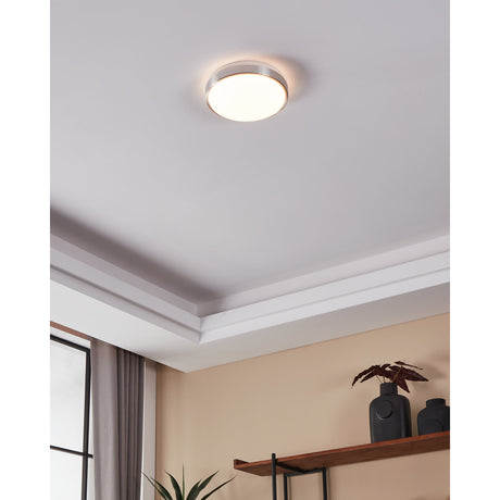 Competa 1 LED flush ceiling light, 24.5cm round white diffuser with satin nickel coloured band, shown illuminated on a ceiling in a room with coving and warm-toned walls.