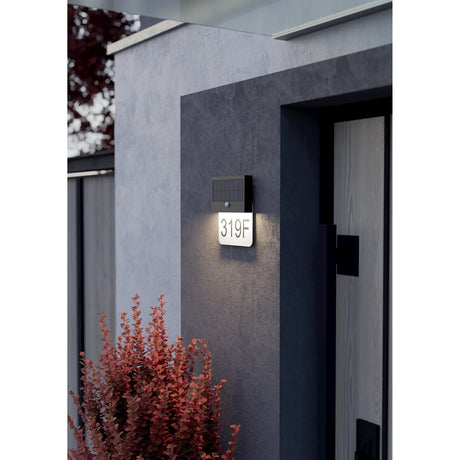 Conettimara solar wall light with motion sensor in black, mounted beside a front door, illuminating an opal house number plate "319F" in warm white light.