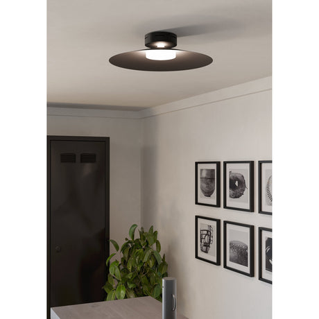 Conomavilla Smart LED Flush Ceiling Light in black, with a round steel canopy and wide flat disc around a white diffuser, shown lit on a hallway ceiling near framed artwork.