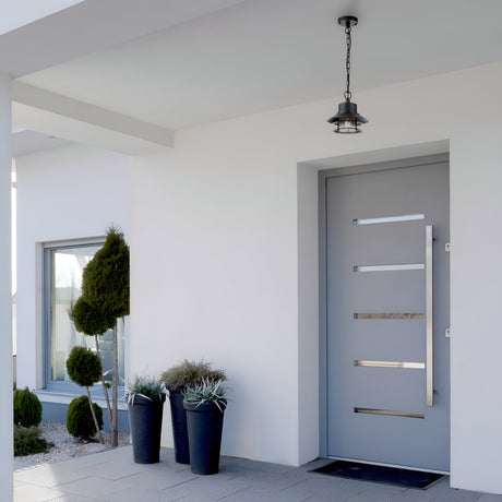 A modern house entrance with a light gray door featuring horizontal glass inserts showcases Danish design. By the door, three black planters filled with greenery are accompanied by the Copenhagen Outdoor 1 Light Chain Pendant in black overhead. A large window on the left enhances the coastal charm.