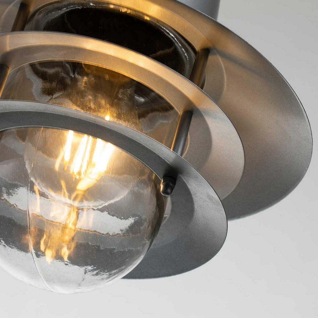 Close-up of the Copenhagen Outdoor 1 Light Chain Pendant in Silver, showcasing a glowing bulb centered within its layered, metallic circular design. The light diffuses through the clear, textured glass, casting a warm and ambient glow.