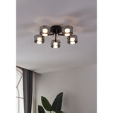 Copillos 5 Light Ceiling Light in black steel with smoked glass cylindrical shades, mounted flush on a white ceiling in a room with sheer curtains and a leafy plant below.