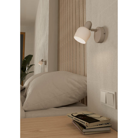 Corato Wall Spotlight in sand and satin nickel, shown beside a bed with an adjustable white glass shade illuminating the wall above a nightstand with books.