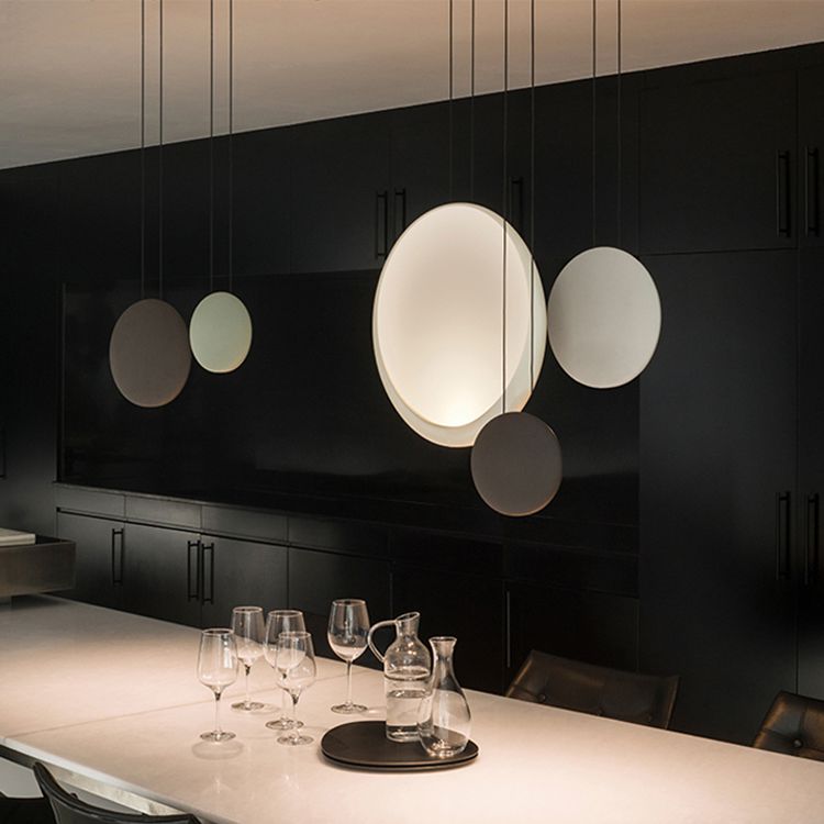 The contemporary dining room features a sophisticated black theme, accentuated by the Cosmos LED Small Pendant Light that emits a soft glow above the white table. Prioritizing energy efficiency, four empty wine glasses and a clear carafe are elegantly arranged on a dark tray, completing the refined atmosphere.