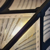 The Chapel 1 Light Outdoor Flush Light - Black features a textured frosted glass panel with diagonal etched lines, beautifully illuminated from behind. Its die-cast aluminium black metal framework adds geometric detail, creating an abstract design with warm light glowing through the glass—ideal for use as an IP44-rated outdoor flush light.