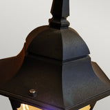 Close-up of the Chapel 1 Light Outdoor Pedestal Lantern in black, showcasing its durable die-cast aluminum structure and geometric design. This striking outdoor lighting piece features a curved top and pointed finial, emitting a warm glow. Set against a soft gradient of light gray, it enhances the elegant charm of any outdoor space.