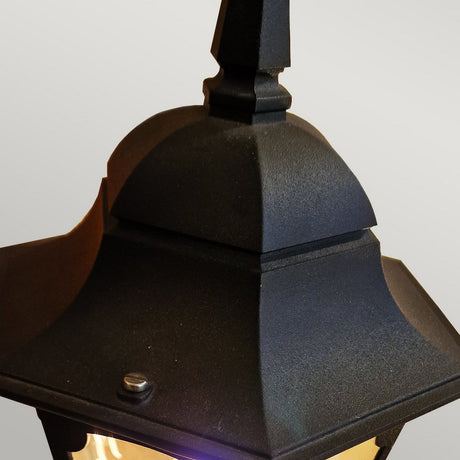 Close-up of the Chapel 1 Light Outdoor Pedestal Lantern in black, showcasing its durable die-cast aluminum structure and geometric design. This striking outdoor lighting piece features a curved top and pointed finial, emitting a warm glow. Set against a soft gradient of light gray, it enhances the elegant charm of any outdoor space.