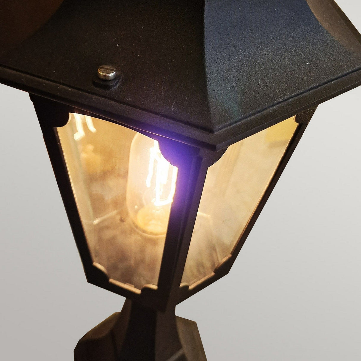 The Chapel 1 Light Outdoor Pedestal Lantern in Black stands against a plain background, its glowing light bulb enhancing the vintage style. Made from durable die-cast aluminum, the lantern's frame and glass panels offer an elegant and classic appearance.