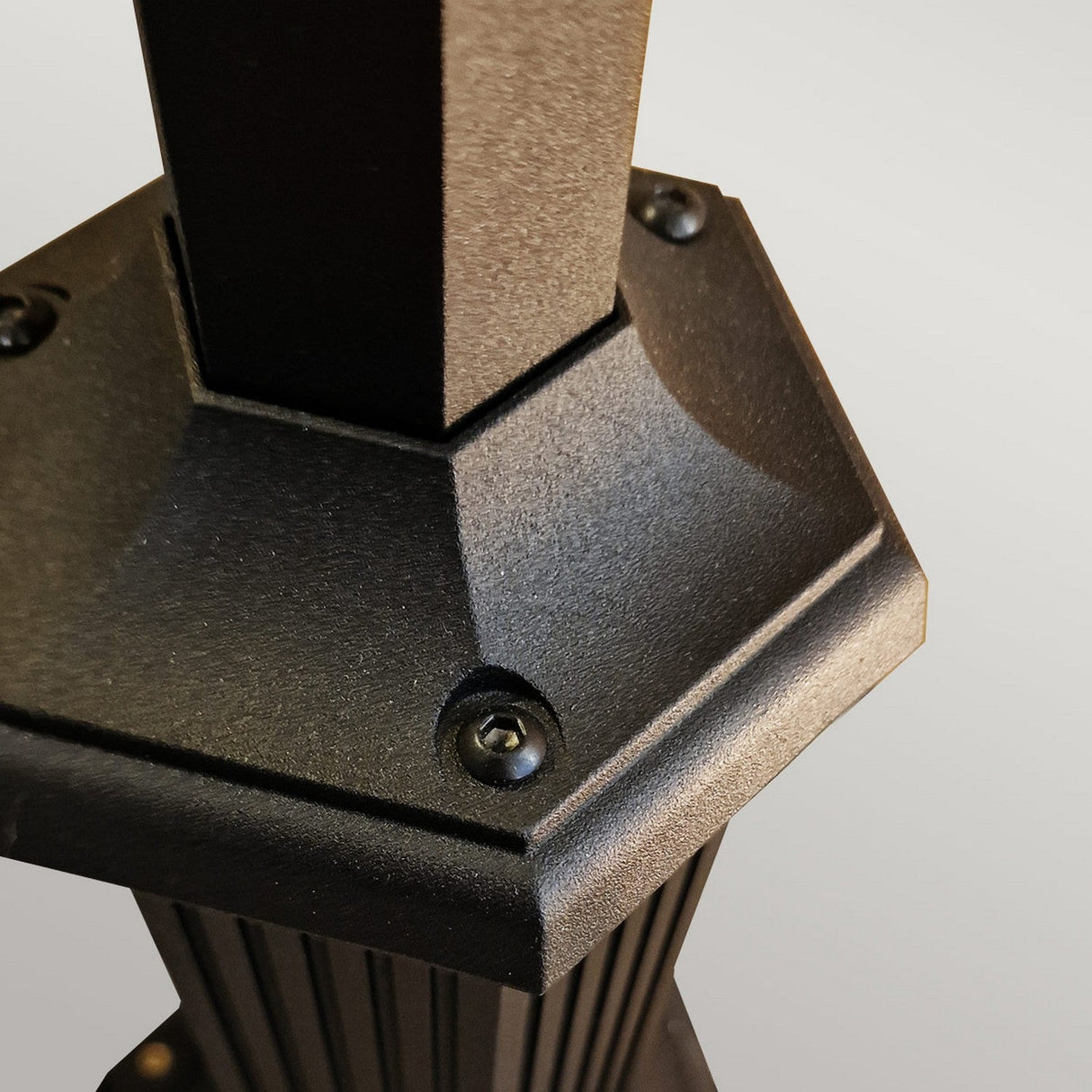 Close-up of the hexagonal base in die-cast aluminum of the Chapel 1 Light Lamp Post - Black, featuring a vertical post secured by screws at the corners. The matte finish evokes sleek outdoor lighting, set against a soft gray backdrop.