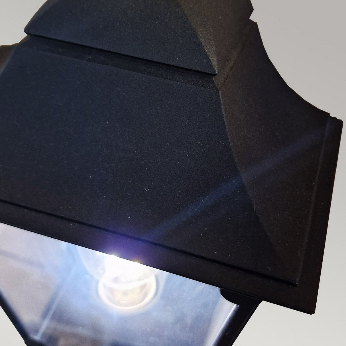 A close-up of the illuminated Chapel-1 1 Light Outdoor Wall Lantern in black, crafted from die-cast aluminum. Its geometric design and bright light stand out against a soft gradient of light gray, emphasizing its IP44 rating and sturdy construction.