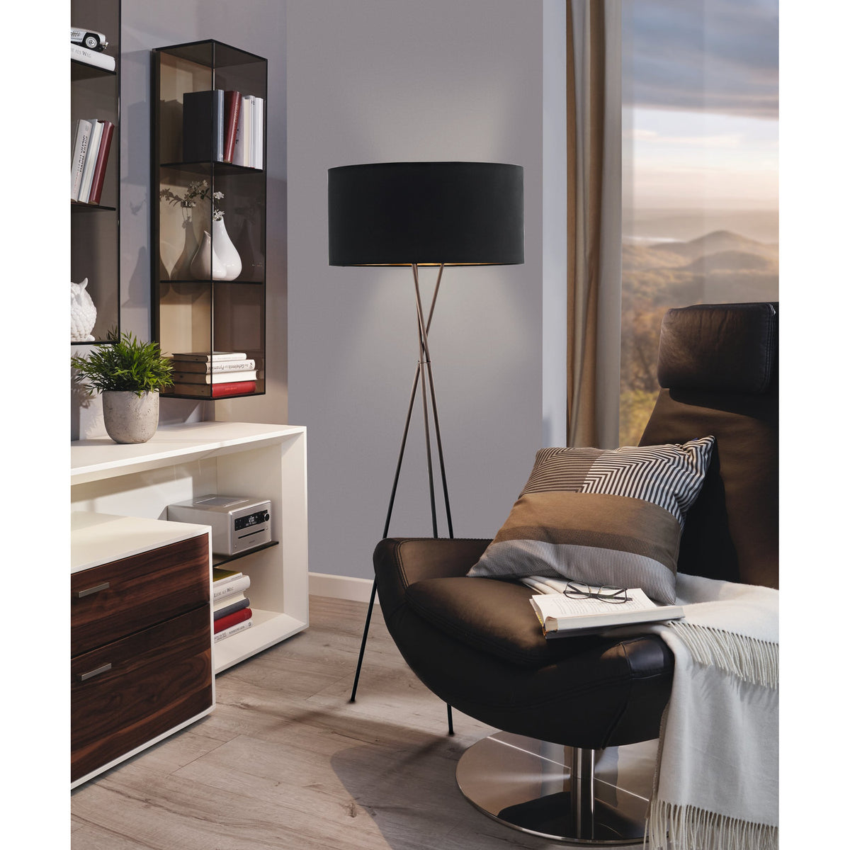 Crelvane Floor Lamp in black, tripod steel legs with a wide fabric drum shade, shown beside an armchair in a living room setting.