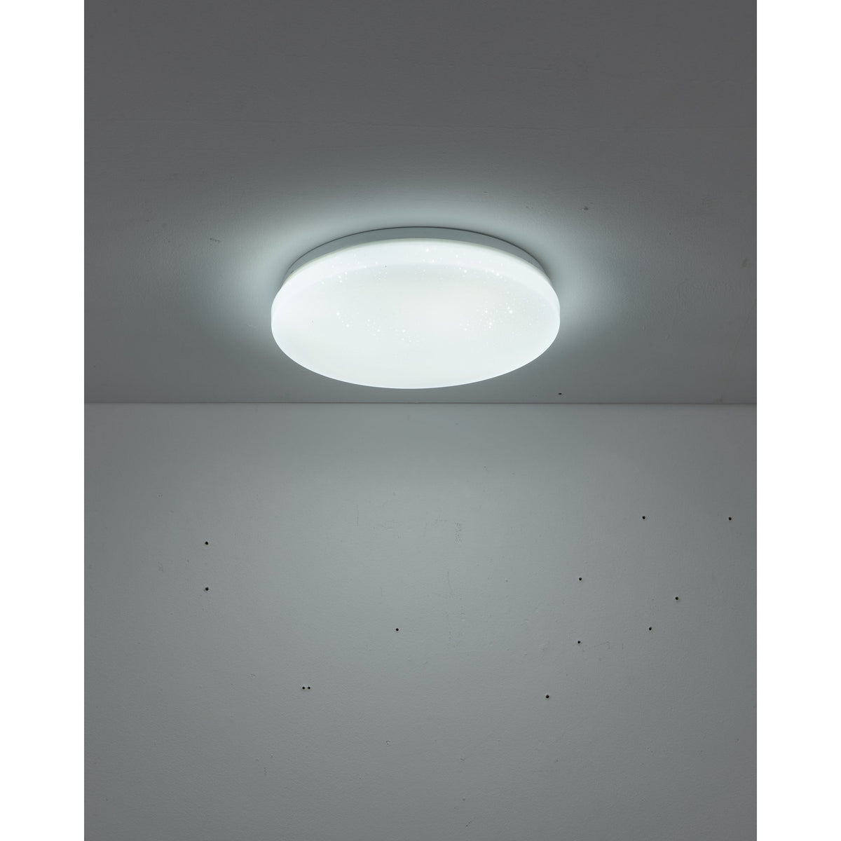 Crelvette round flush LED ceiling light with white steel body and crystal-effect plastic diffuser, illuminated on a grey ceiling in a room setting.