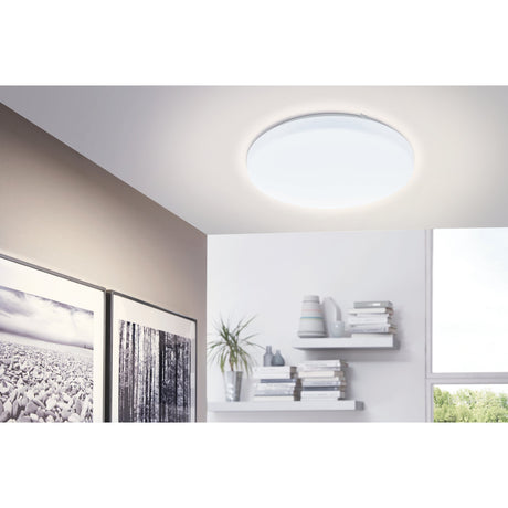 Crelvette round LED flush ceiling light, 33cm, white steel body with opal plastic diffuser, shown glowing warm white on a hallway ceiling near shelves and artwork.