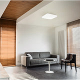 Crelvette square LED ceiling panel light in white, 33 x 33cm with rounded diffuser, mounted on a living room ceiling above a grey sofa and black chair.