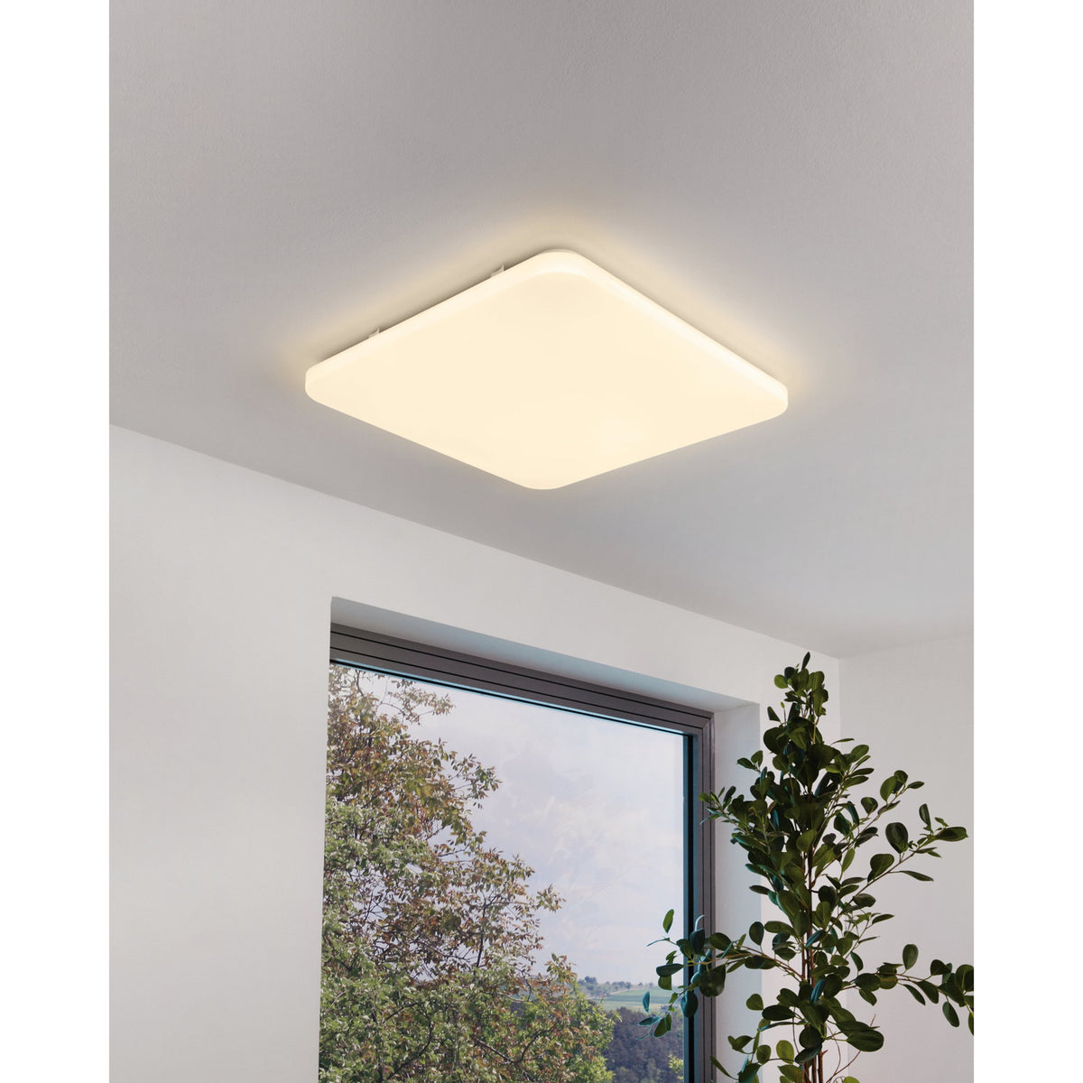 Crelvette 53 x 53cm square LED ceiling panel light in white, mounted flush on a bedroom ceiling with warm white glow, shown near a window and plant.