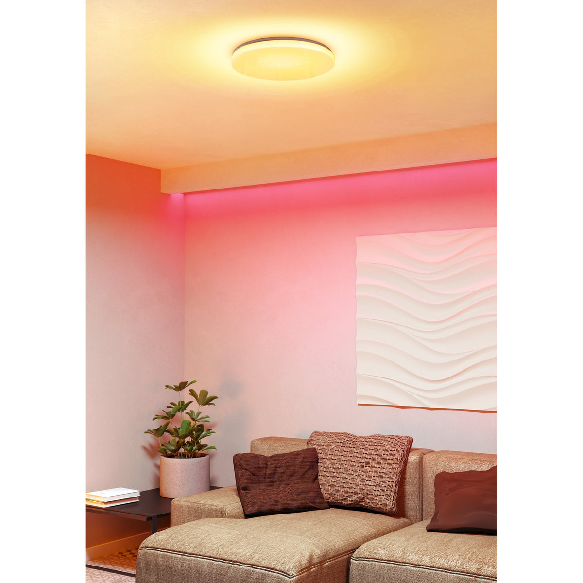 Crelvette round LED flush ceiling light in white, shown glowing warm with pink RGB accent lighting along the wall in a living room with a sofa and cushions.