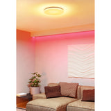 Crelvette round LED flush ceiling light in white, shown glowing warm with pink RGB accent lighting along the wall in a living room with a sofa and cushions.