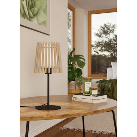 Crolenne Table Lamp in black with slim steel stem, round base, and tapered pale wooden slat shade, shown on a wooden coffee table in a living room setting.