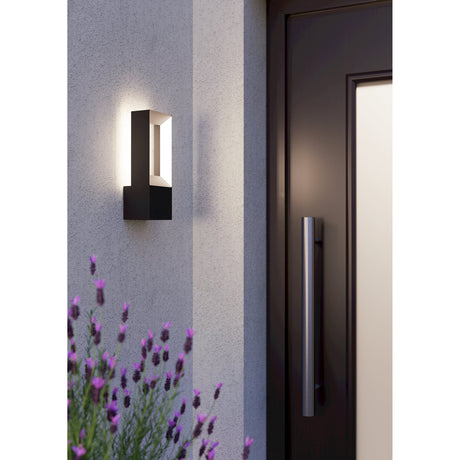 Cronvena LED Outdoor Wall Light in black aluminium, rectangular open frame over a solid base, mounted beside a front door and glowing softly against a grey wall.