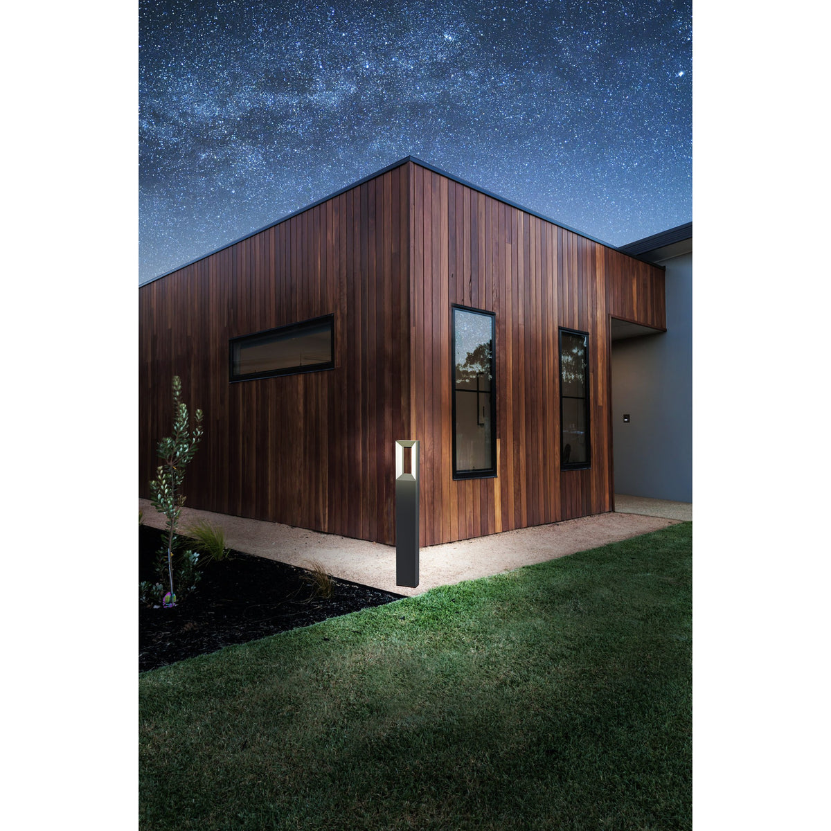 Cronvena outdoor LED floor lamp in black, a slim bollard-style aluminium post with rectangular head, shown illuminating a gravel path beside a timber-clad house at night.
