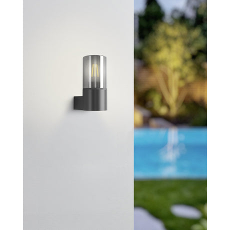 Cropani Outdoor Wall Light in black with a cylindrical smoked glass shade and square side bracket, shown on a white wall beside a blurred garden and pool.
