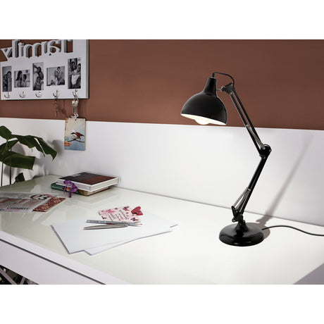Crosmanne Table Lamp in gloss black steel with white-lined dome shade, jointed arm and round base, shown on a desk beside papers and a plant.