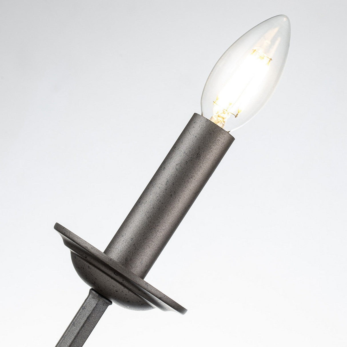 A close-up of a lit candle-shaped LED bulb on the Crown 2 Light Wall Light - Graphite reveals its timeless design, which emits a warm glow and contrasts beautifully with the graphite finish against a light background.