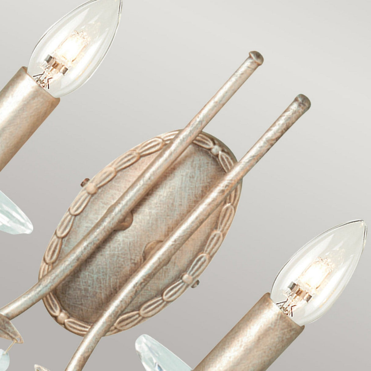 A detailed view of the Christina 2 Light Wall Light - Silver/Gold Patina showcases two decorative bulbs on either side. The fixture features an oval base with slender rods finished in a stylish Silver and Gold Patina, adorned with crystal drops. The elegantly shaped bulbs resemble candle flames.