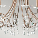 A close-up view of the Christina 8 Light Chandelier in Silver/Gold Patina showcases its ornate design, featuring curved metallic arms decorated with dangling prisms. The soft, neutral grey background highlights the chandelier's elegant design, making it an ideal choice for formal dining rooms.