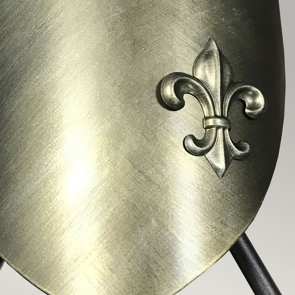 The Crusader Wall Light in Burnished Bronze features a close-up of a metal shield with a brushed finish, showcasing a raised fleur-de-lis emblem on the right side. The medieval design is highlighted by touches of Burnished Bronze against the neutral gray background.