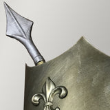 The image showcases a close-up of a metallic shield corner adorned with a medieval fleur-de-lis emblem. An arrow is positioned at the top edge, highlighting its burnished bronze finish reminiscent of a Crusader Wall Light's subtle illumination, set against a plain light gray background.