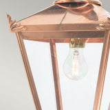Close-up of the Chelsea Outdoor 1 Light Down Wall Lantern - Copper, featuring clear glass panels that showcase a visible light bulb. This timeless piece of exterior decor lighting is adorned with elegant decorative metalwork at the corners. Its weathered look adds character against the soft gray background.