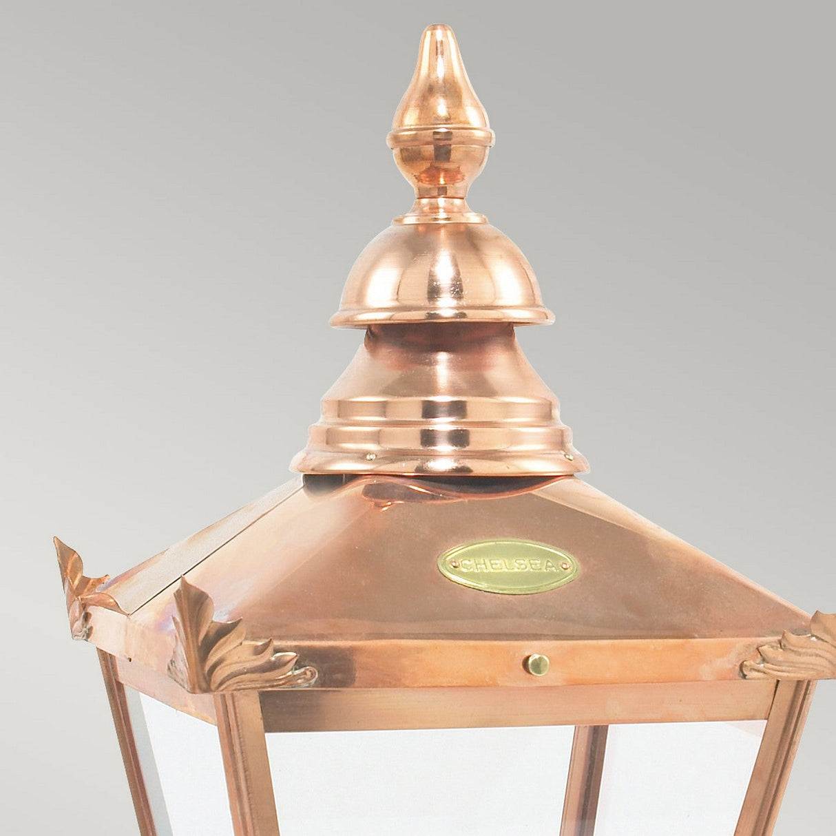 A close-up of the Chelsea 1 Light Pedestal Lantern - Copper highlights the polished finial and an embossed plaque with "Chelsea" on it. The frame features decorative corner elements, making it a striking piece of outdoor decor against the neutral gray background.