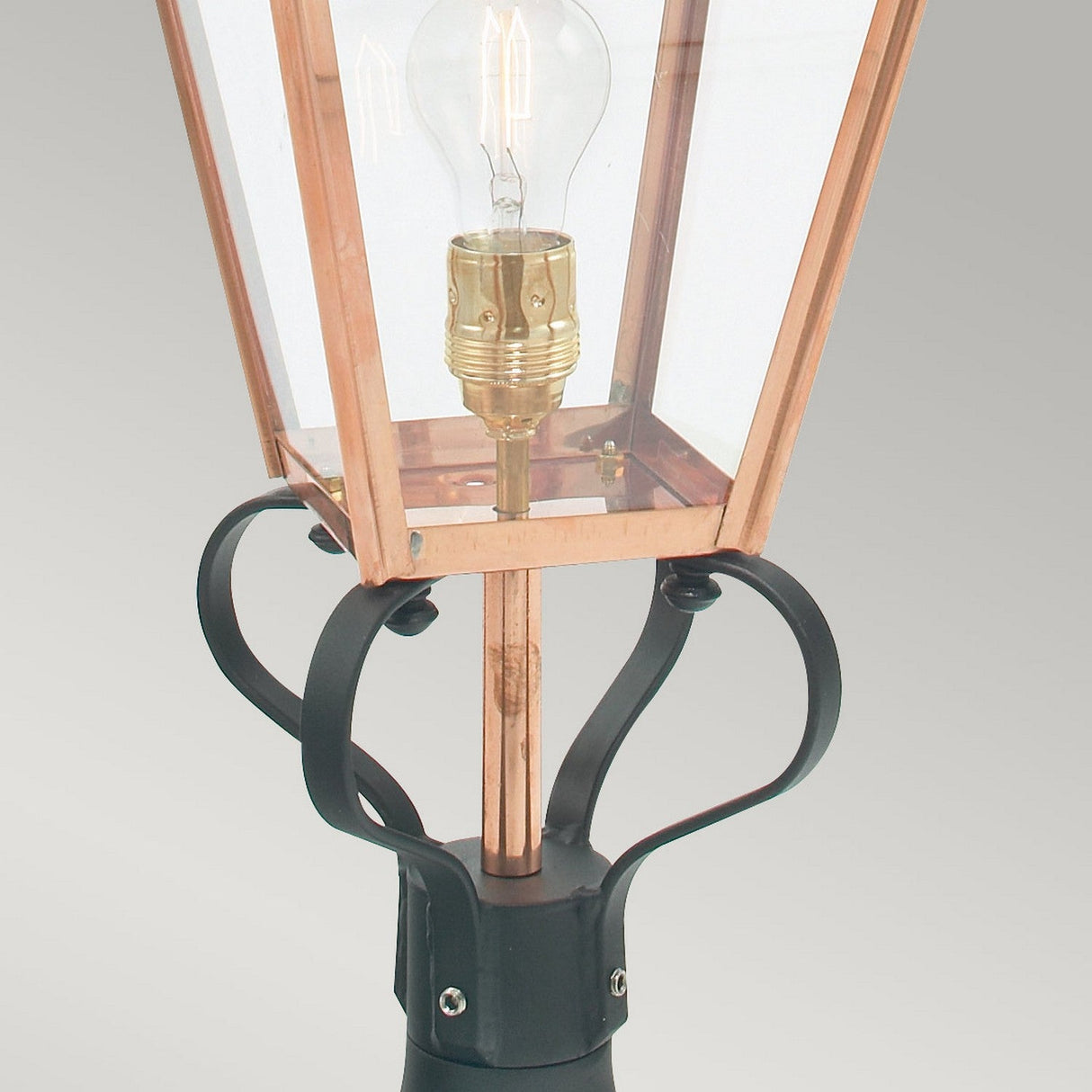A close-up of the Chelsea 1 Light Pedestal Lantern in copper features a transparent glass case that showcases its weathered character. The clear light bulb inside highlights the black metal base with decorative curved designs, making it a standout piece for outdoor decor.