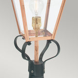 A close-up of the Chelsea 1 Light Pedestal Lantern in copper features a transparent glass case that showcases its weathered character. The clear light bulb inside highlights the black metal base with decorative curved designs, making it a standout piece for outdoor decor.