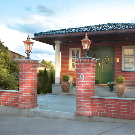 A well-maintained brick house with a green door is elegantly adorned by Chelsea 1 Light Pedestal Lanterns in copper on brick pillars flanking the entrance. The garden, showcasing tasteful outdoor decor with potted plants, sits under a sky that’s partly cloudy.