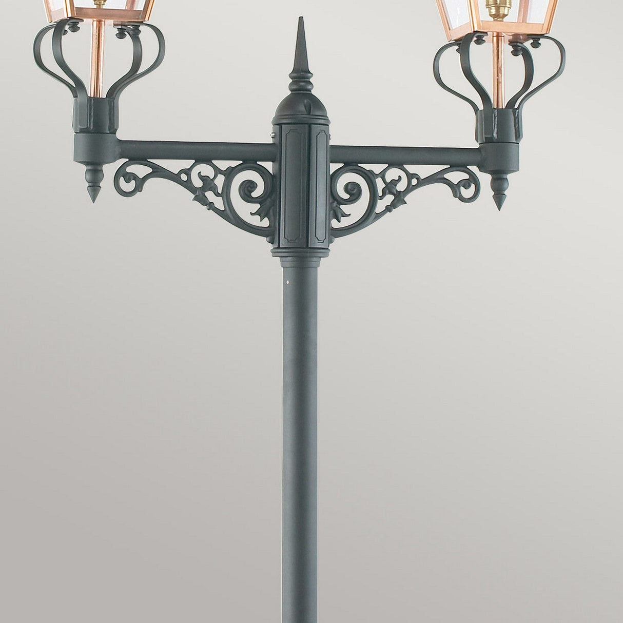 The Chelsea Twin Head Lamp Post - Copper & Black showcases a black, ornate design with two lanterns and decorative scrollwork beneath each light. Its angular lighting shines through clear panels, highlighting intricate details in dazzling Copper & Black tones against a neutral backdrop.